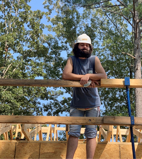 A young man wearing a hard hat stands at a house under construction.