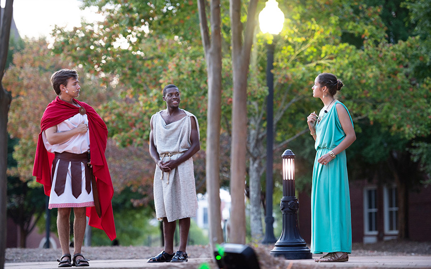 Three students dressed in Greek/Roman attire perform “The Braggart Soldier” outdoors with trees and a lamp post in the background.