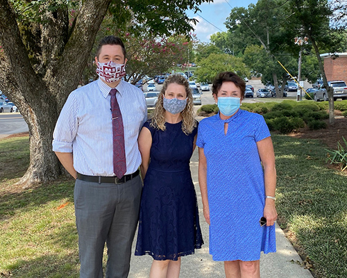 A man and two women stand together outside with trees, grass and a parking area in the background.