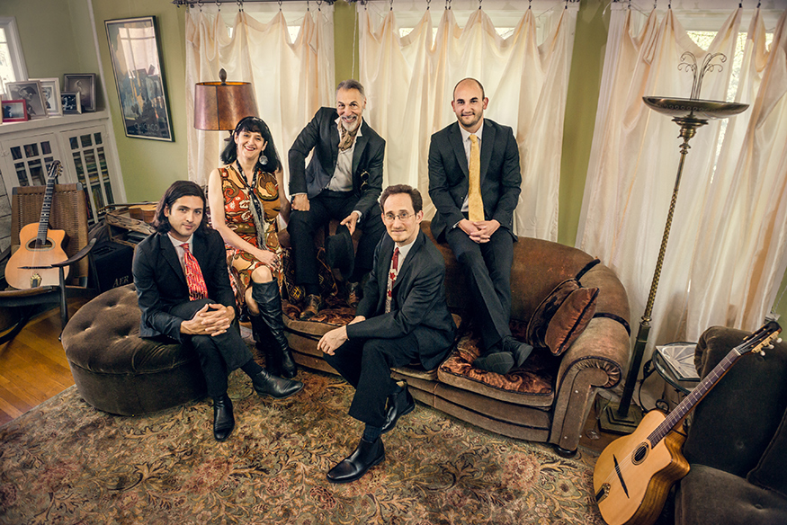 Members of the Hot Club of San Francisco are pictured seated on a couch near windows with white drapes.