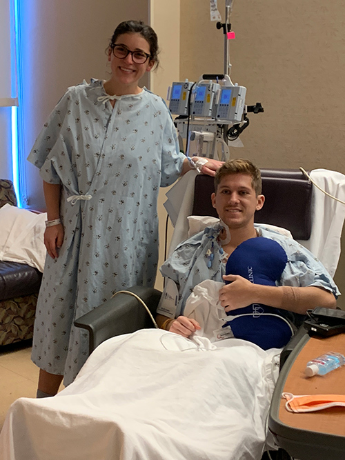 A woman in a hospital gown stands next to a man in a hospital bed. Both smile at the camera.