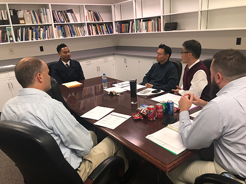 A student sits at a large table with other men in a room with shelves of books.