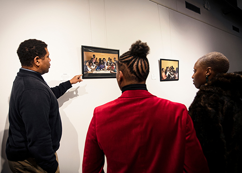 African-American individuals look on as an African-American man points at a photograph on the wall.