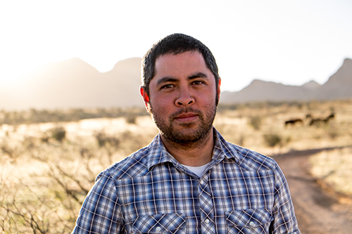 A man stares at the camera while standing in a desert with mountains in the distance.