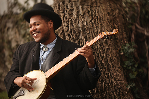 Jerron “Blind Boy” Paxton, seated next to a tree and smiling while holding a banjo.