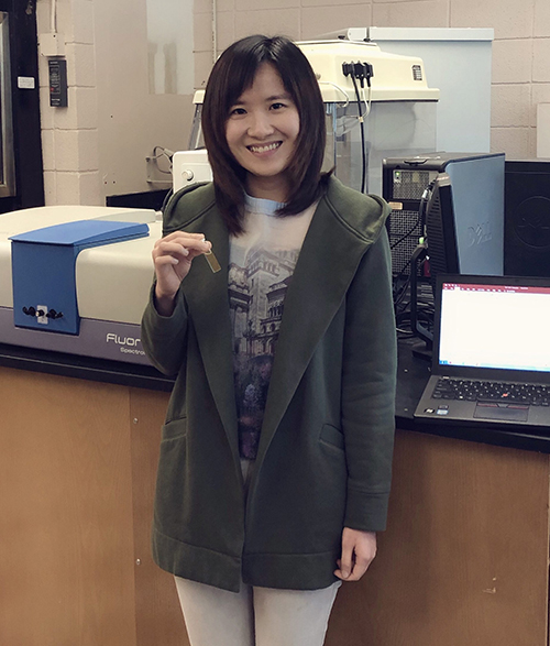 Joanna Xu holds a fluorescence cuvette sample. Xu’s MSU lab contains a fluorometer (background) with which she acquired fluorescence spectra.