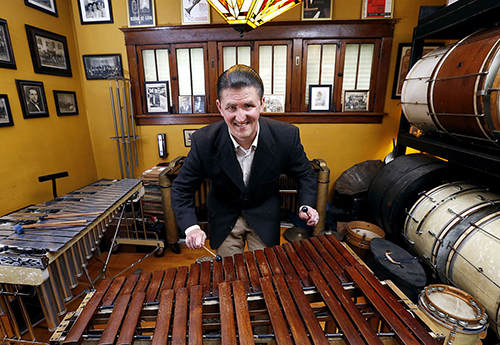 Josh Duffee smiles for the camera while standing in a yellow room with xylophones and drums