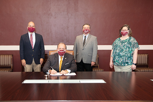 MSU President Mark E. Keenum is seated at a large table while MSU Interim Dean of Libraries Tommy Anderson, MSU Associate Dean of Libraries Stephen Cunetto and MSU Libraries Automations System Manager Anita Winger stand behind him in front of a maroon wall.