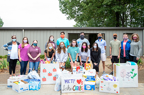 A group of 15 people wearing masks stand by boxes of supplies to be donated