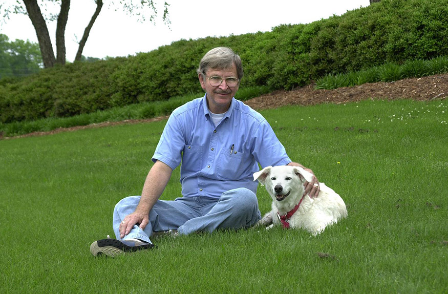 Malcolm Mabry Jr. sits on grass with a white dog, Lulu, beside him