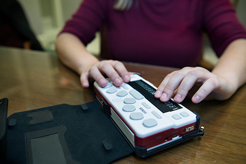 A student in a maroon shirt demonstrates her maroon and white Braille tablet resting on a brown table.