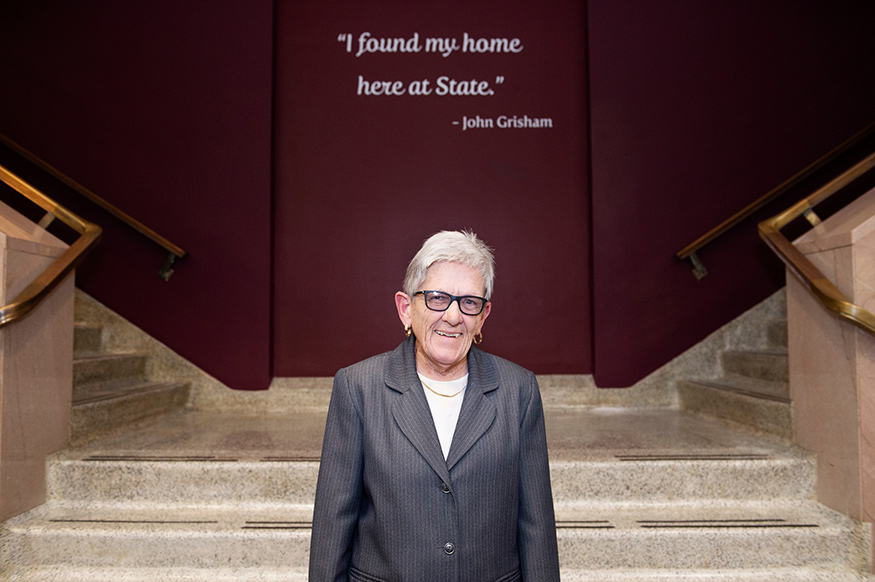 A woman stands in front of a staircase and a maroon wall that includes a quote from John Grisham “I found my home here at State.”