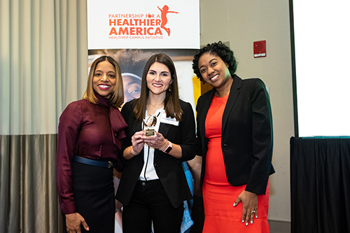 Taylor VanDyk, center, registered dietician and health and wellness educator for Mississippi State’s Department of Health Promotion and Wellness, accepts an apple-shaped award recognizing the university’s three-year efforts to fulfill 23 health and wellness guidelines developed by the Partnership for a Healthier America. Award presenters were Kimberly O’Shields, far left, PHA senior manager of partnerships, and Andrea Muscadin, far right, PHA vice president of partnerships. (Photo submitted)
