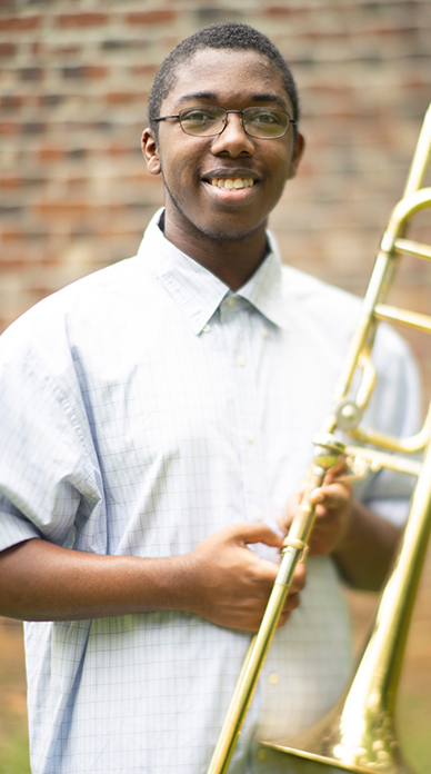 Deontrae Peyton smiles for the camera and holds a gold trombone while standing in front of a brick wall.