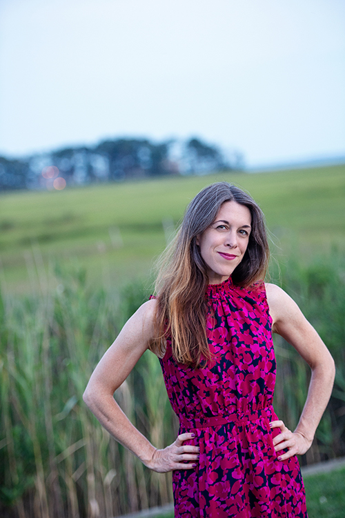 Catherine Pierce stands with her hands on her hips while wearing a bright pink dress in a grassy field.