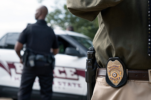 A close-up shot of an officer's badge with another officer and patrol car in the background