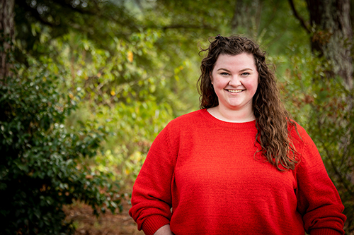 Portrait of Reagan Poston wearing a bright orange shirt while standing in a forest