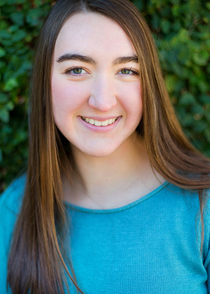 Hannah Scheaffer wears a bright blue shirt and smiles for the camera while standing in front of green bushes.