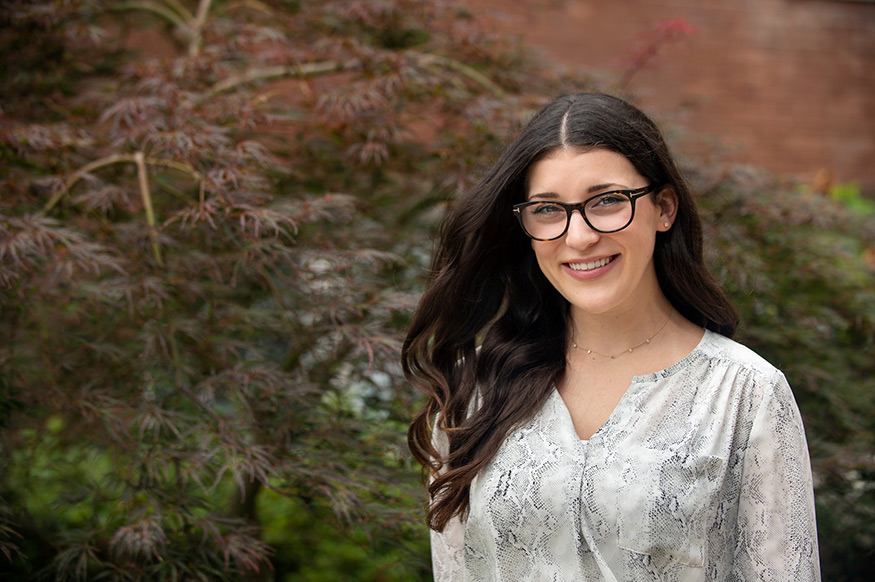 A young woman with long brown hair and glasses smiles with trees in the background