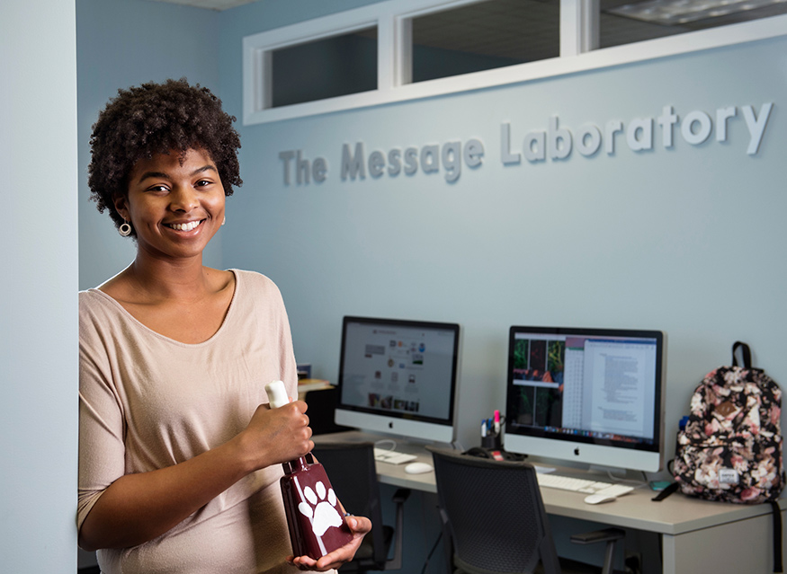 Ciarra Smith holds a cowbell as she stands in front of two computers with "The Message Laboratory" signage above.