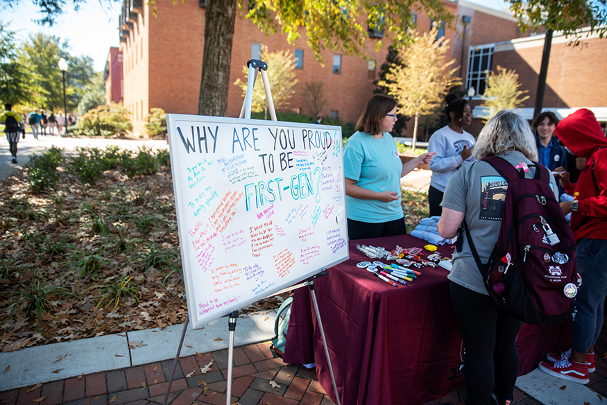 Students talk at an outdoor table next to a white board that reads "Why are you proud to be first-gen?"