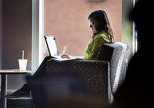 A student seated near a large window glances at a notebook while typing on a laptop.