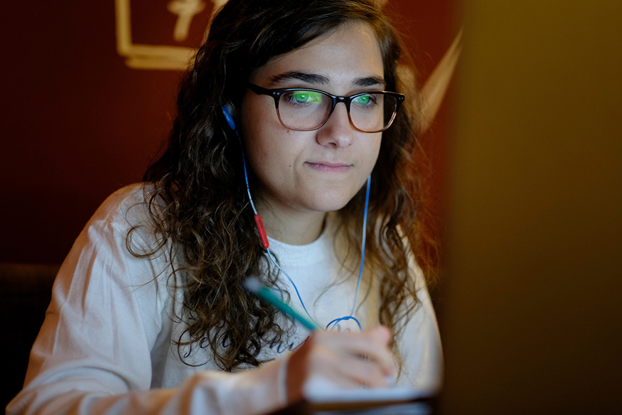 A female student with long dark hair and glasses looks at her laptop