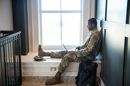 A student in military fatigues work on his laptop in front of a window on campus.