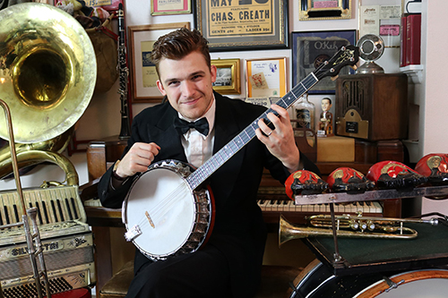 TJ Müller, pictured surrounded by musical instruments, stares at the camera while holding a banjo