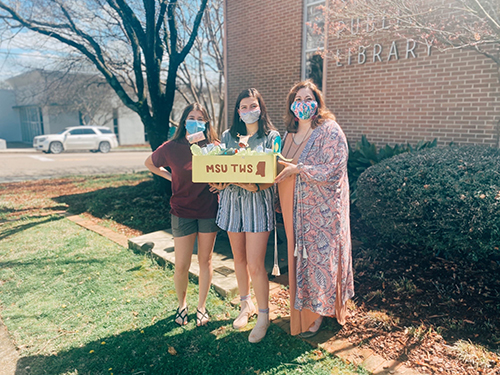 Three masked women stand outside of Starkville Public Library while holding a box labeled with "MSU TWS" and maroon image of the state of Mississippi