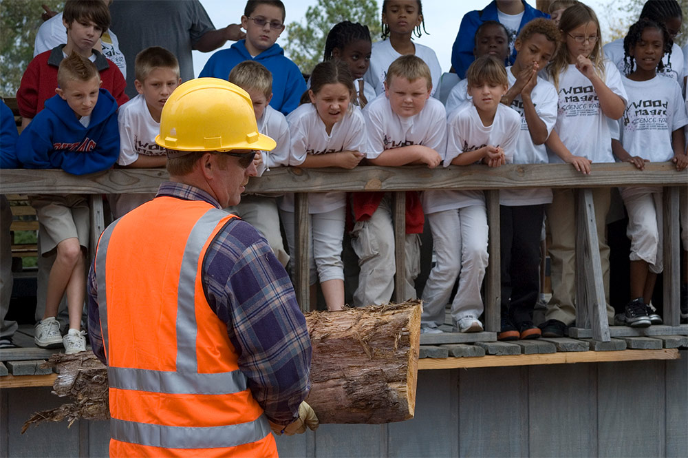 a man wearing a hard hat shows a piece of a log to school children at the Wood Magic Science Fair.