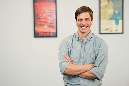 Joey Thompson smiles with his arms crossed while standing in front of a wall with two movie posters on it.