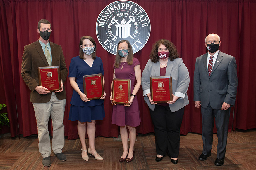 Four faculty members holding awards stand with MSU Provost David Shaw in front of the MSU seal.