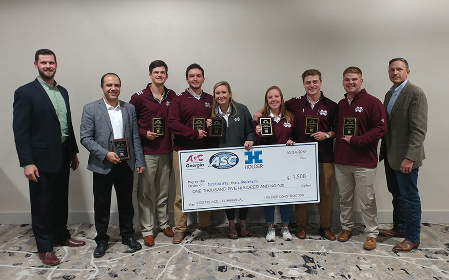 A group of nine students and faculty hold a large check and individual plaques.