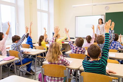 Students raise their hands while their teacher stands at the front of the classroom.