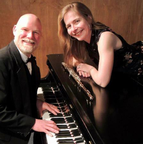 Jeff Barnhart smiles while seated at a piano his wife Anne Barnhart is laying on while smiling