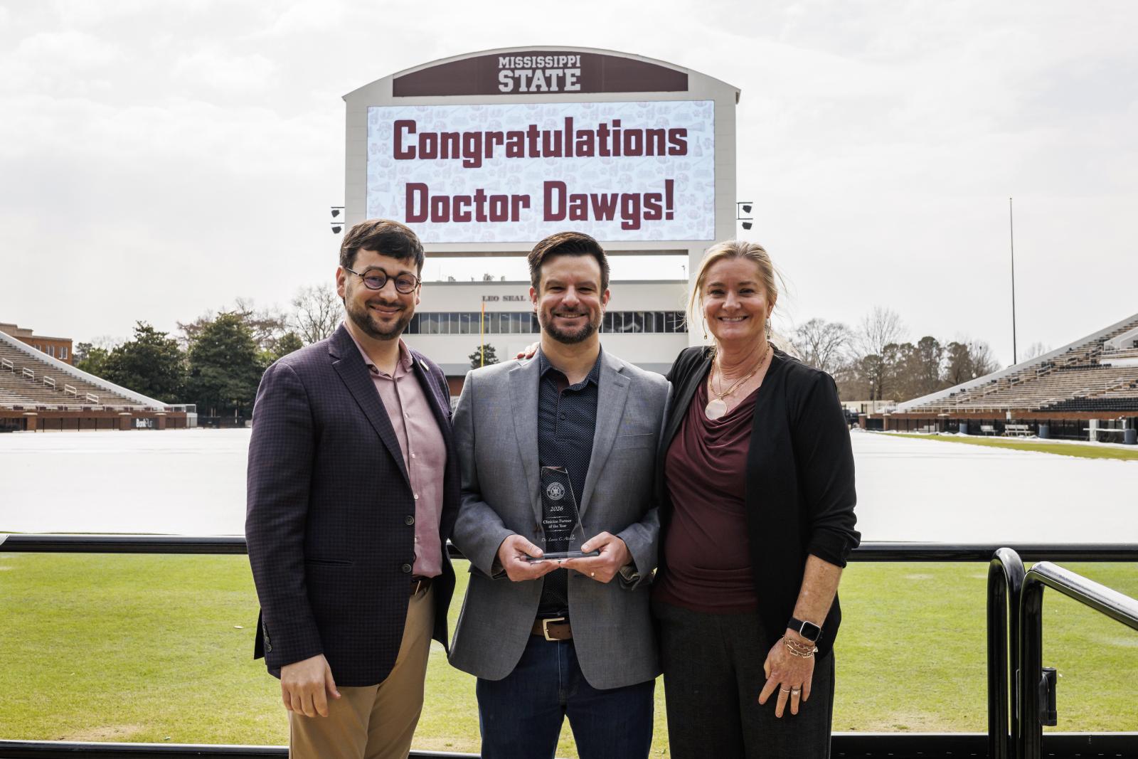 Two men and a woman stand with an award at a football stadium
