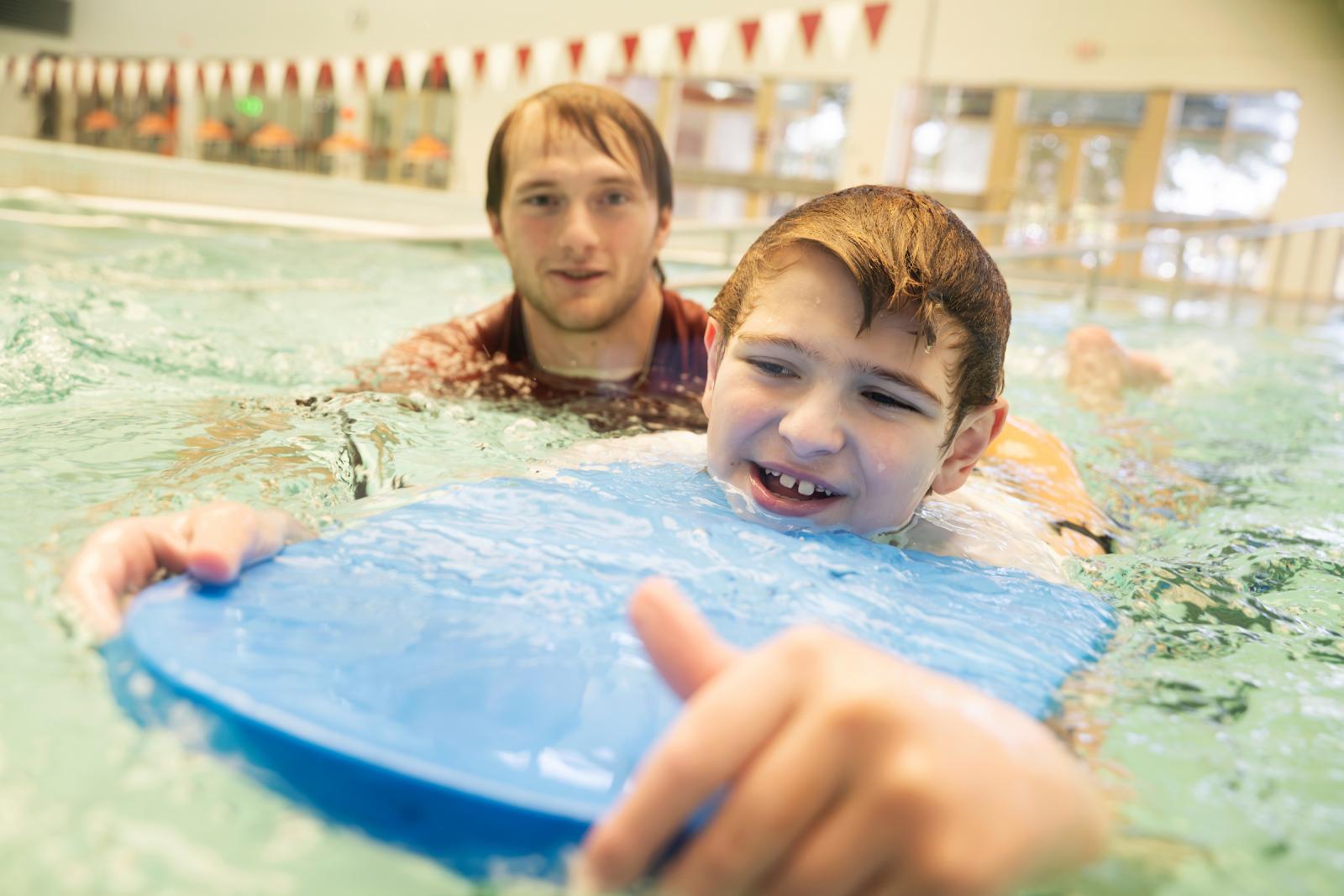 swim instructor teaching a young boy to swim in pool with kickboard