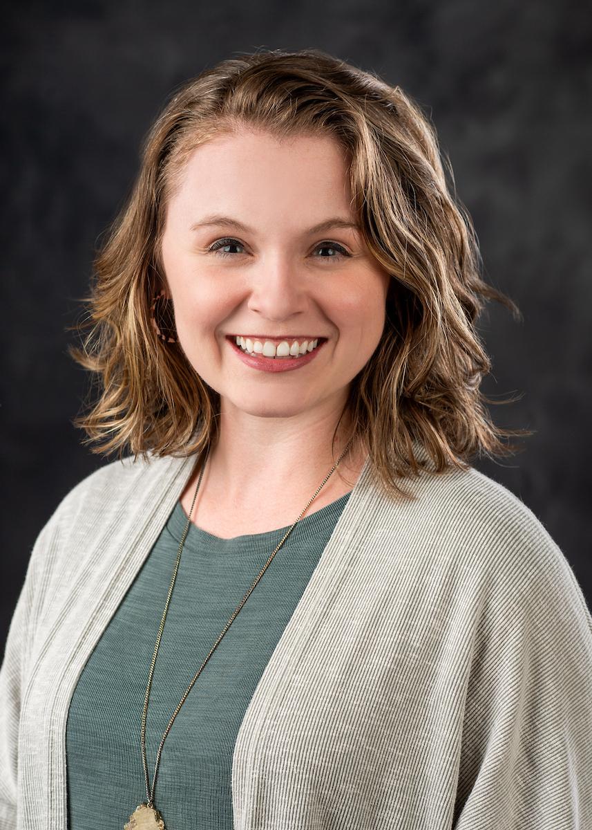 Headshot of Kenna Vowell in green shirt and grey sweater on grey backgound