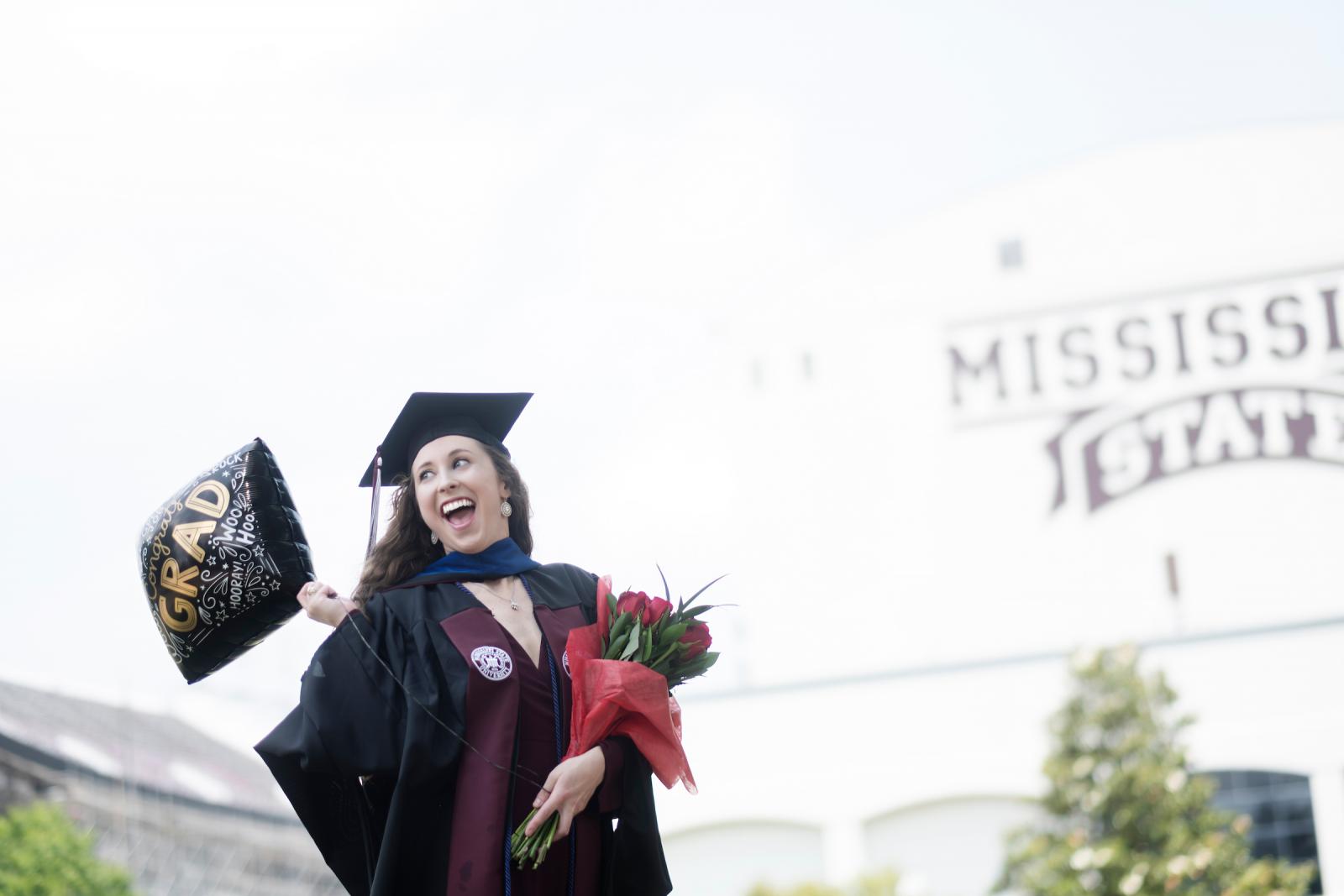 a woman holding graduation balloons and flowers in front of MSU stadium
