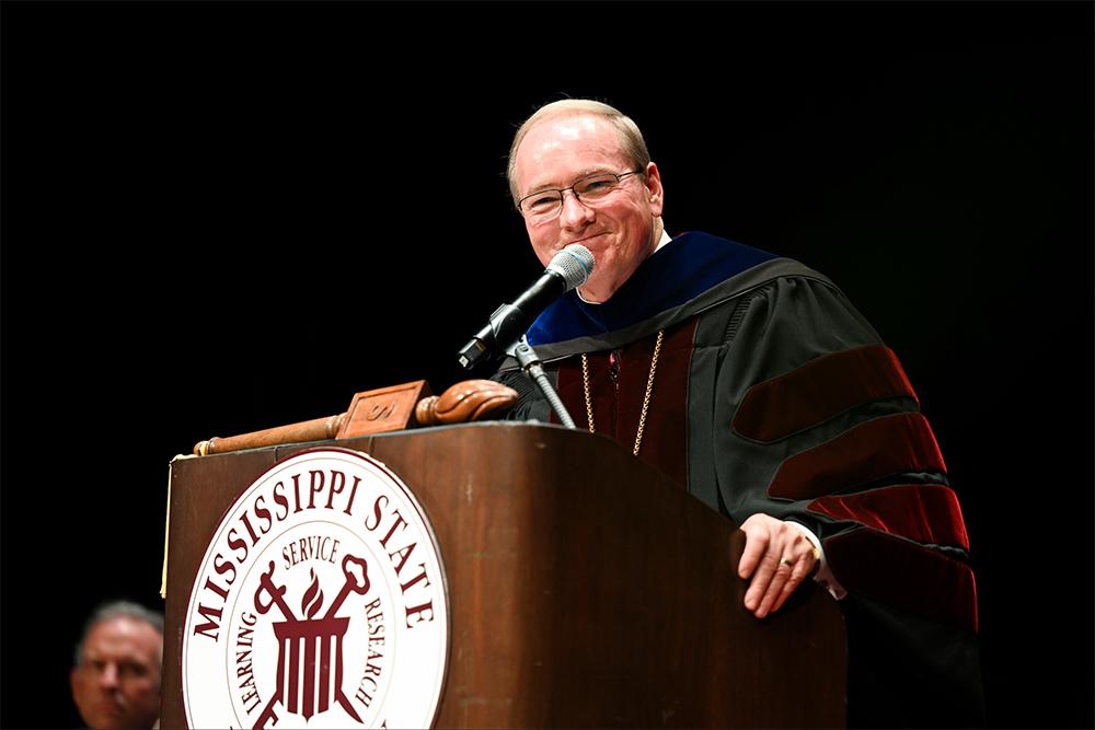 MSU President Mark E. Keenum stands at a speaking podium while wearing graduation regalia