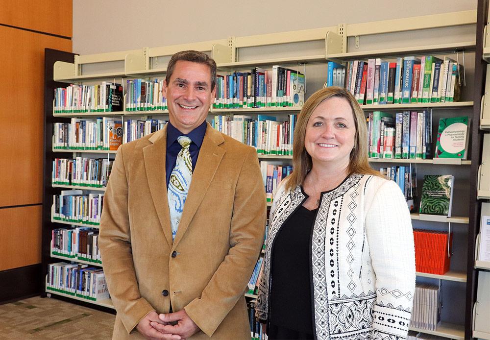 A man and woman stand in a library or office with a full bookshelf in the background