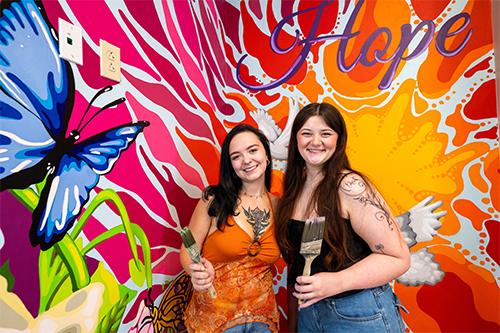 Two art majors stand by a brightly colored mural they painted