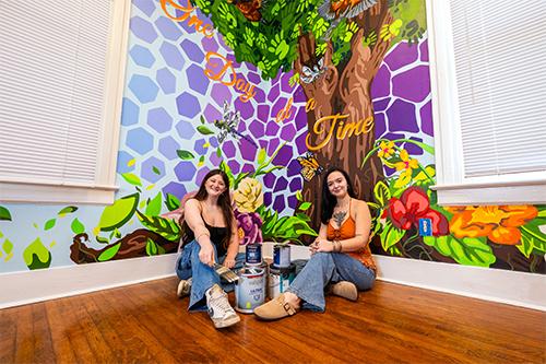 Two art students sit by a brightly colored wall mural