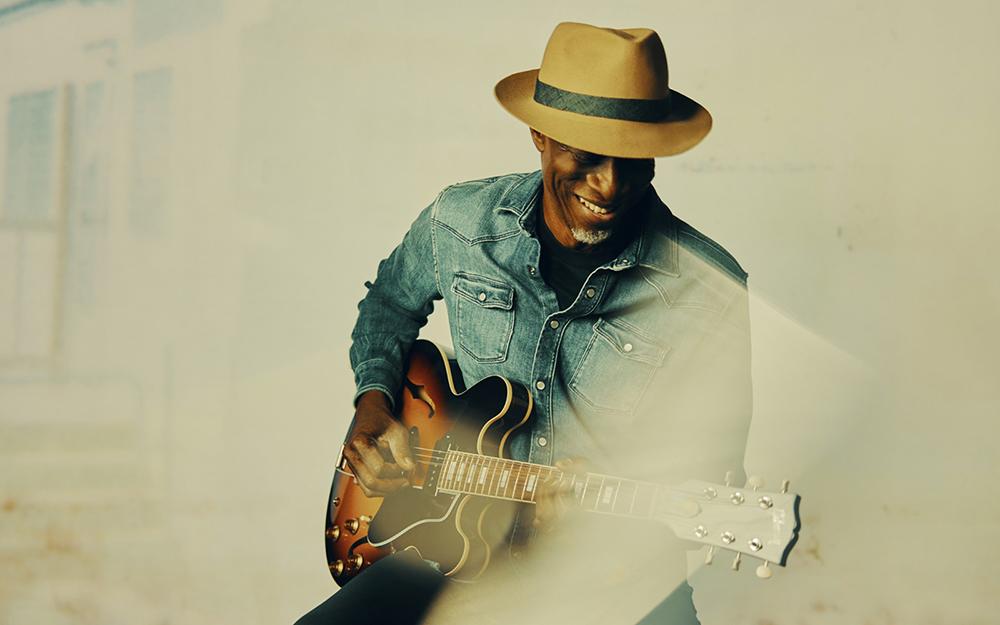A promotional photo of blues musician Keb' Mo' while he plays guitar.