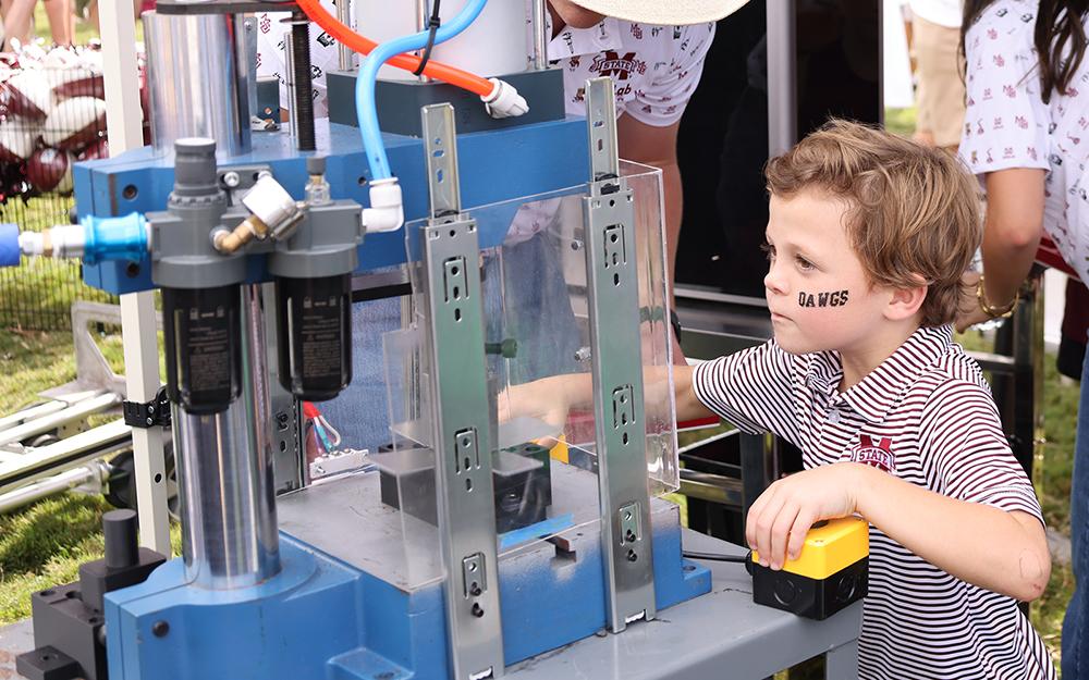 Mississippi State IDEELab faculty member Collin Mullins teaches a young Bulldog fan the stamping process with the hydraulic press of making a cowbell during pregame festivities at the Junction on Saturday, Sept. 26, 2026. The IDEELab helped children make 209 cowbells. (Photo by Kristen Polk)